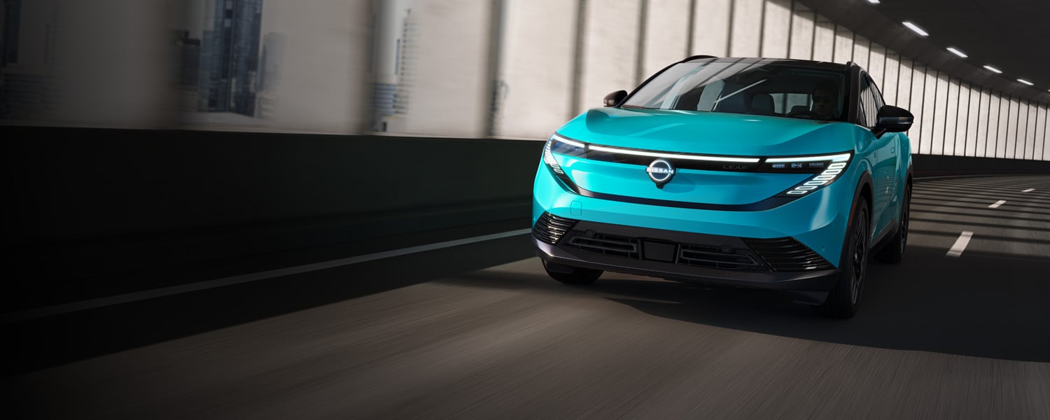 A turquoise Nissan Leaf electric vehicle driving through a modern tunnel, with sunlight streaming through vertical windows on the side.
