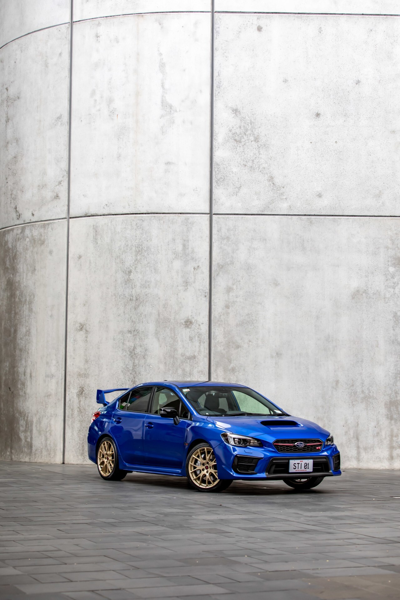 Blue Subaru WRX STI parked in front of a tall concrete wall with gold wheels and a large rear wing.